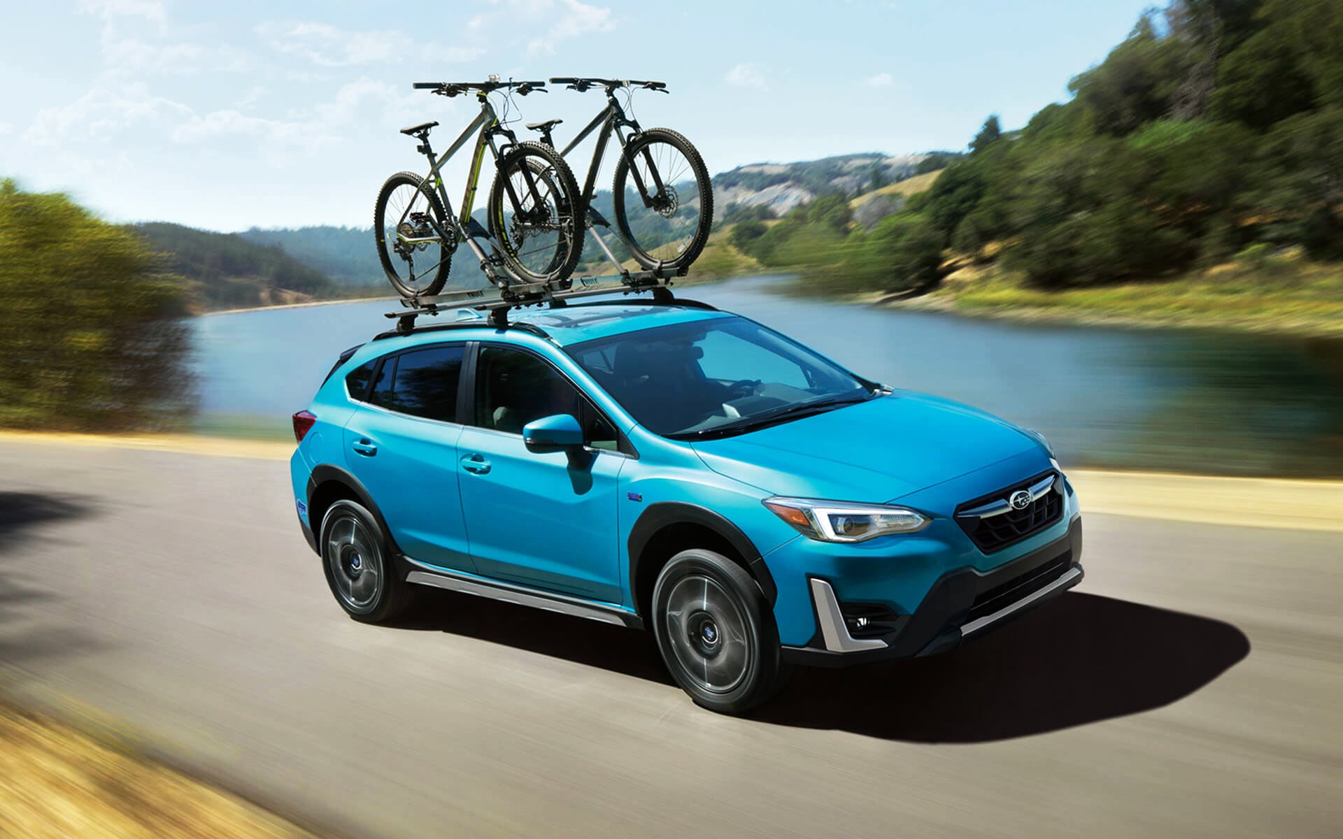 A blue Crosstrek Hybrid with two bicycles on its roof rack driving beside a river | Bergstrom Subaru Green Bay in Green Bay WI